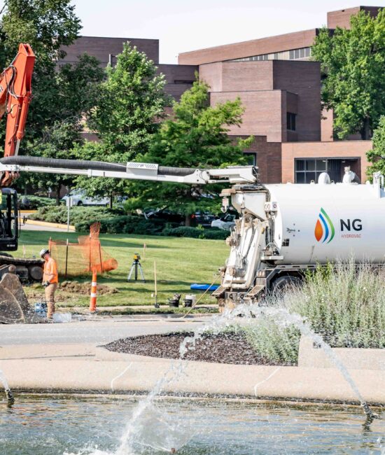 NG Companies hydrovac truck with employee and excavator in front of building with fountain